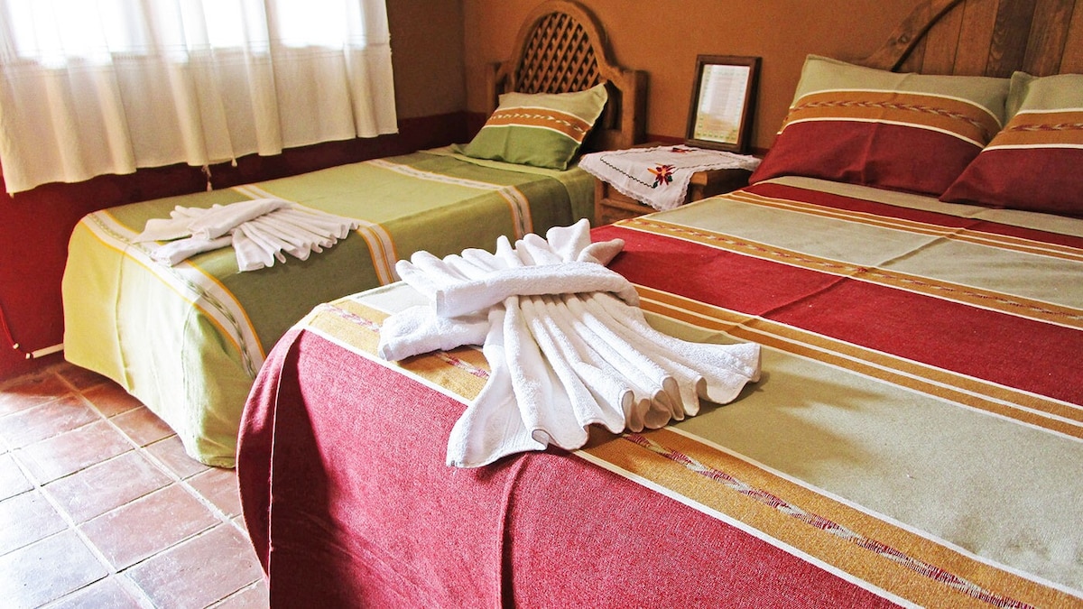 A cozy sleeping area features two spacious beds with colorful woven blankets in green and red hues. Neatly folded white towels are displayed on the beds. Natural light filters through a window covered by sheer curtains, illuminating earthy-toned walls and rustic decor.