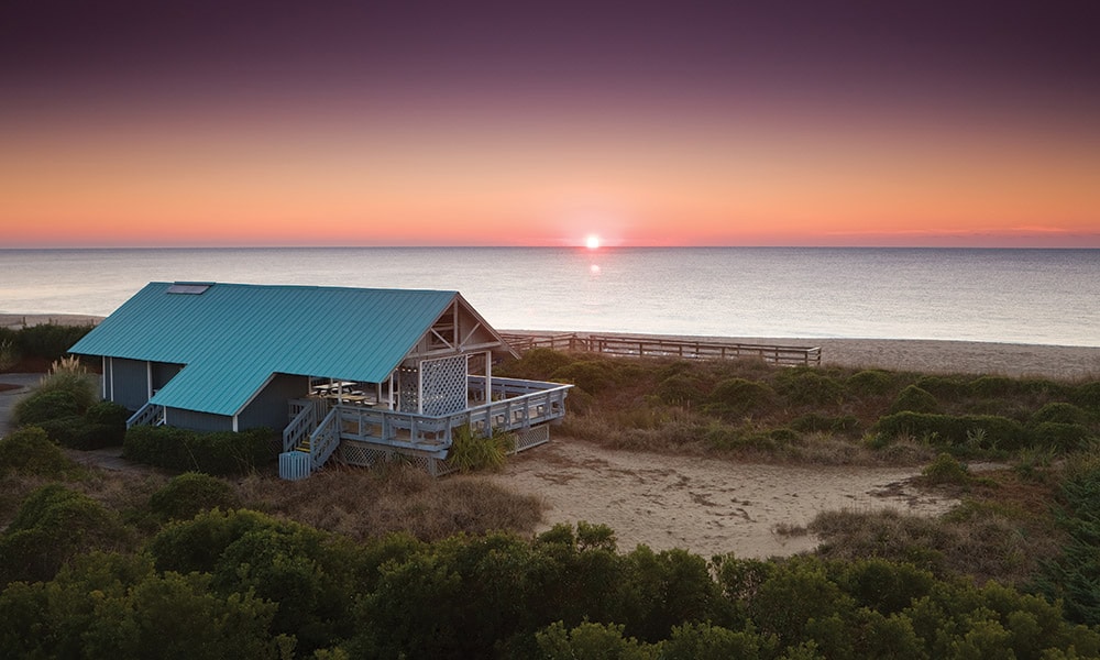 A charming coastal home is situated near the beach, featuring a distinctive blue roof. The sunset casts a warm glow over the horizon, blending orange and purple hues in the sky. Lush greenery surrounds the sandy area leading to the calm ocean waters.