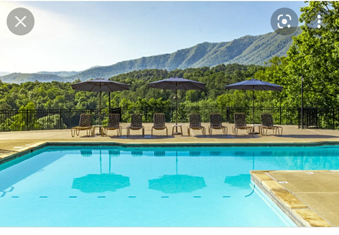 A swimming pool reflects the surrounding mountains under a clear sky. Several lounge chairs are arranged around the pool, each shaded by large umbrellas, providing a tranquil setting for relaxation.