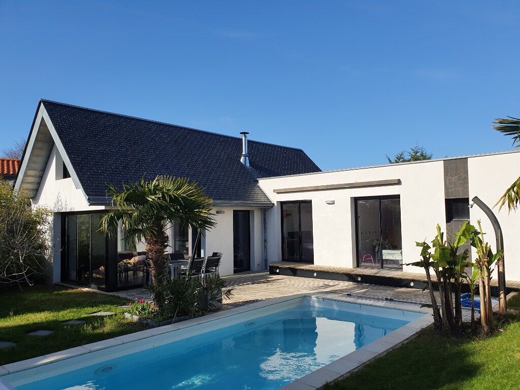 A modern architectural villa is set against a clear blue sky, featuring large glass doors that create an open feel. A lush garden surrounds the space, with a swimming pool at the forefront and palm trees enhancing the serene atmosphere of the outdoor area.