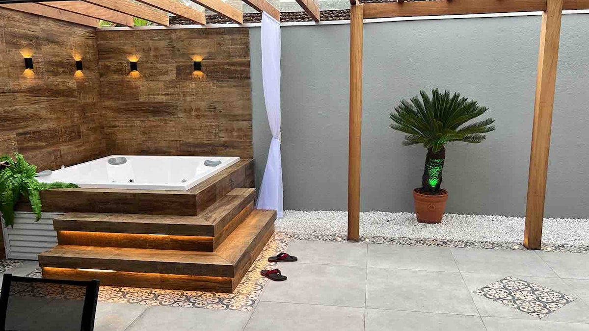 An outdoor area features a wooden deck surrounding a hot tub, illuminated by strategically placed wall lights. A potted plant adds greenery to the space, complemented by light-colored tiles and small stones scattered on the ground. Two pairs of sandals are visible nearby.