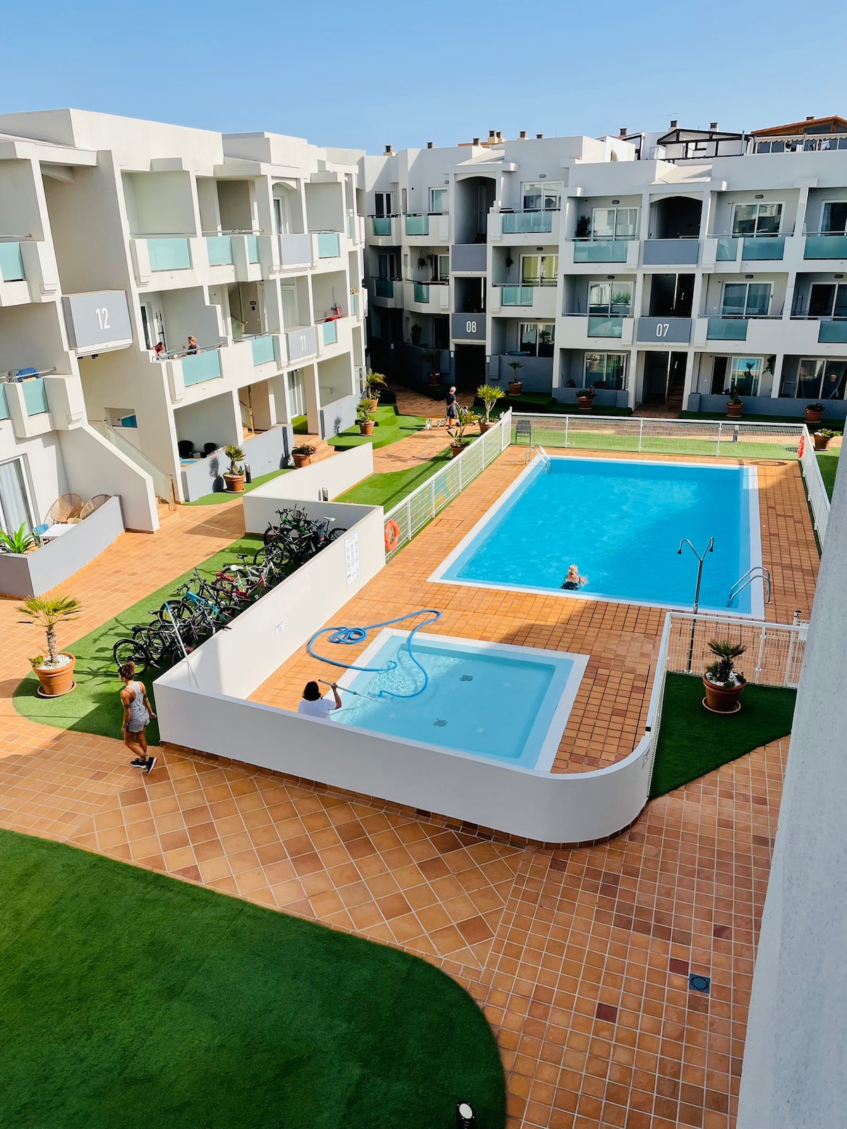 The image captures a serene courtyard featuring two swimming pools surrounded by well-maintained gardens. Sun loungers are placed near the pools, and several people are seen enjoying the space. The area is lined with apartment buildings, showcasing a modern architectural style.
