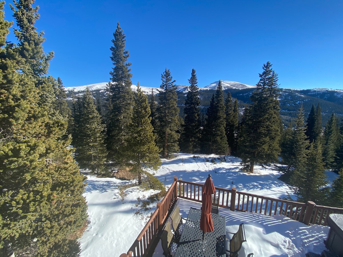 A spacious deck with a table and chairs is visible, surrounded by snow-covered ground and evergreen trees. The background features expansive mountain views under a clear blue sky, creating a serene outdoor setting.