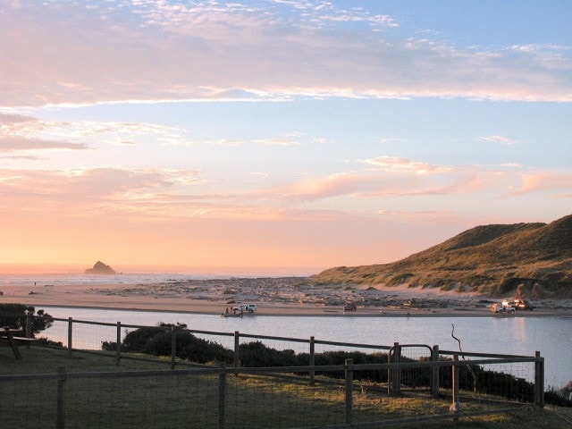 A serene view of the riverfront is presented, with gentle waves reflecting colors of the sunset. Soft hills are visible in the background, complemented by a sandy beach. A wooden fence lines the foreground, framing the picturesque scene that stretches towards the horizon.
