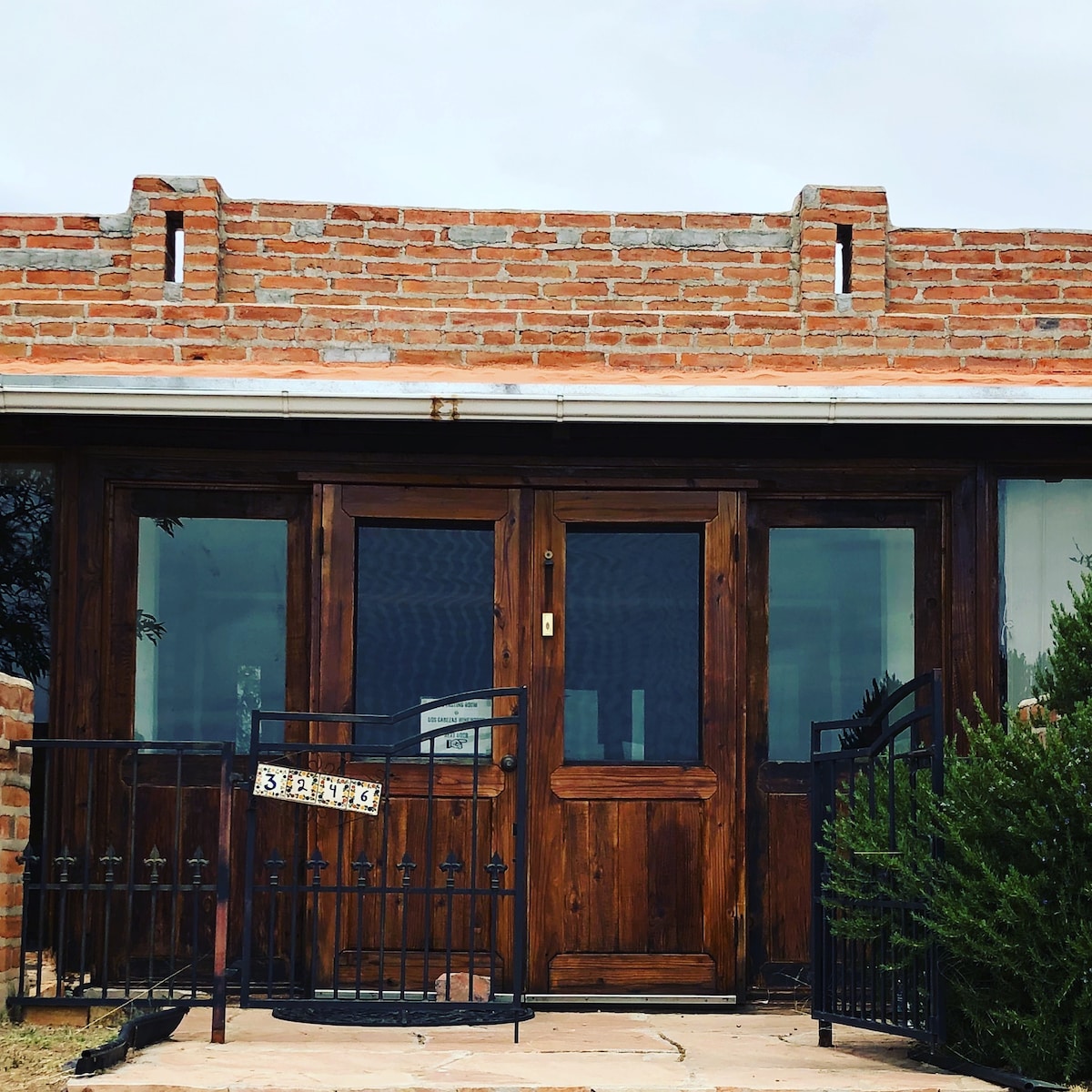A welcoming entrance features two wooden doors framed by brick walls. Decorative wrought iron fencing complements the rustic look. Seasonal greenery can be seen framing the doorway, with a stone path leading to the entrance under a soft gray sky.