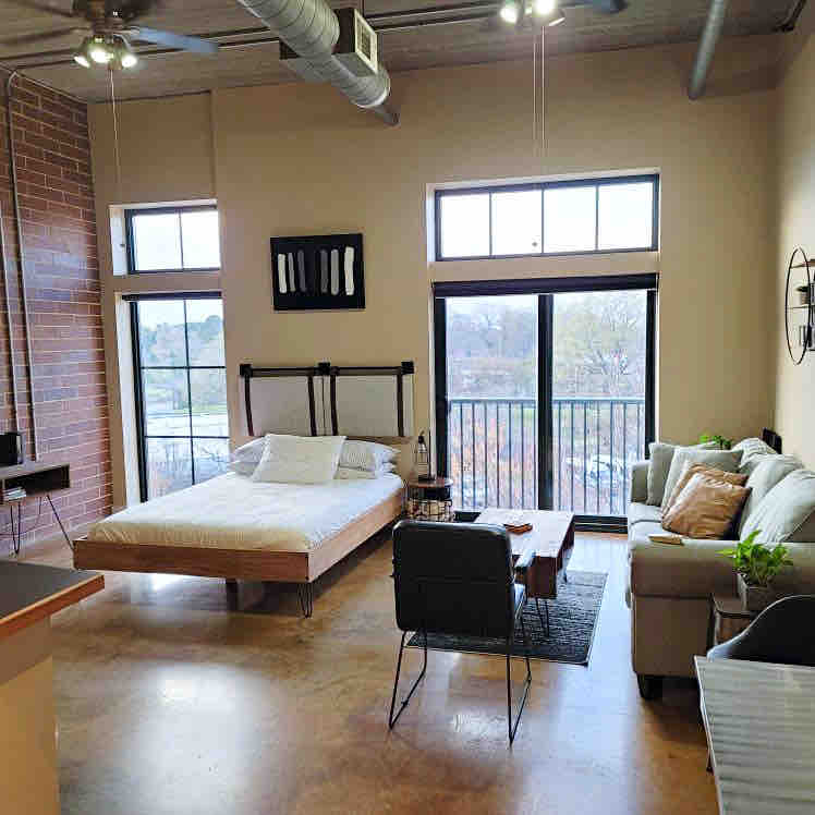 An open studio space features exposed brick walls and concrete floors. A spacious bed sits beside large windows, allowing natural light to fill the room. A seating area with a sofa and armchair is included, complemented by a small coffee table.