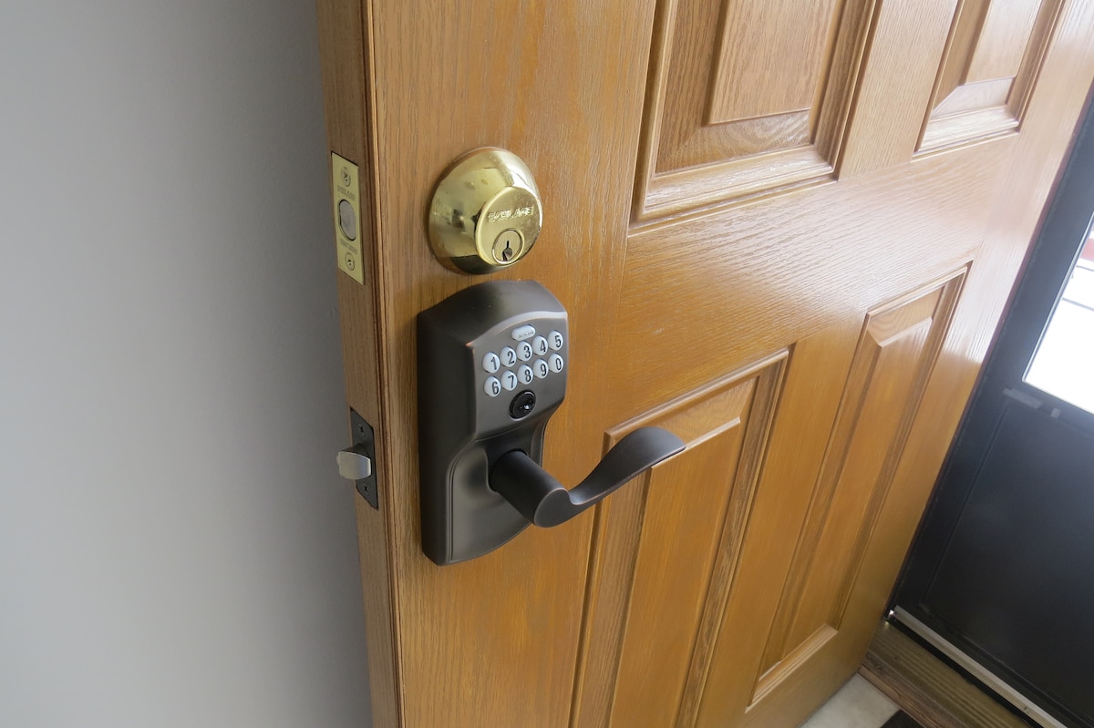 The image features a close-up view of a wooden door with an electronic keypad lock. A round brass doorknob is positioned above the lock, and light reflects off the door's smooth surface. The door frame and adjacent glass panel are also visible.