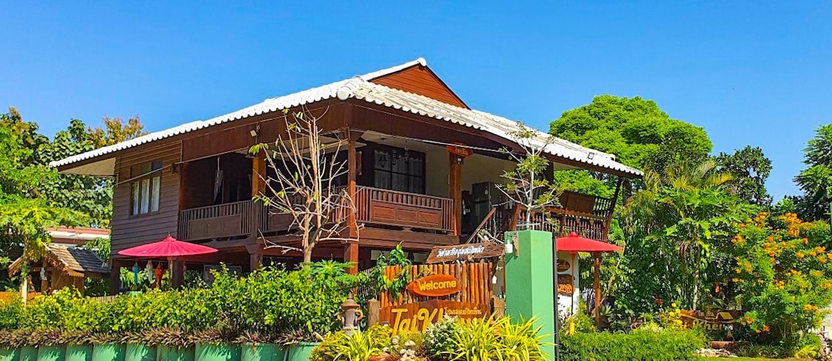 A traditional wooden house features an inviting balcony with an overhanging roof. Surrounding greenery and vibrant flowers enhance the outdoor area, while red umbrellas provide shade. The bright blue sky adds to the welcoming atmosphere of the property.