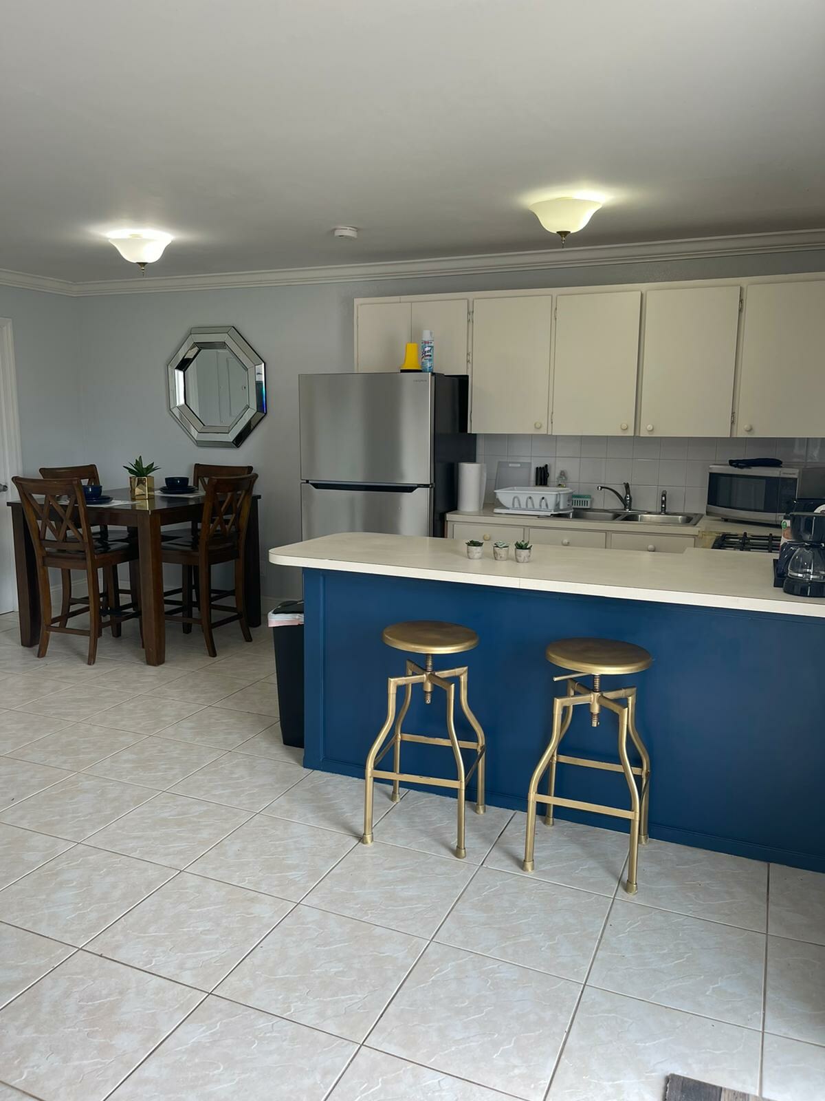 A spacious kitchen area features a blue accent wall, complemented by light-colored cabinetry. A round dining table with four wooden chairs is present, alongside a stainless steel refrigerator and modern appliances. Two gold bar stools are situated at the kitchen counter.