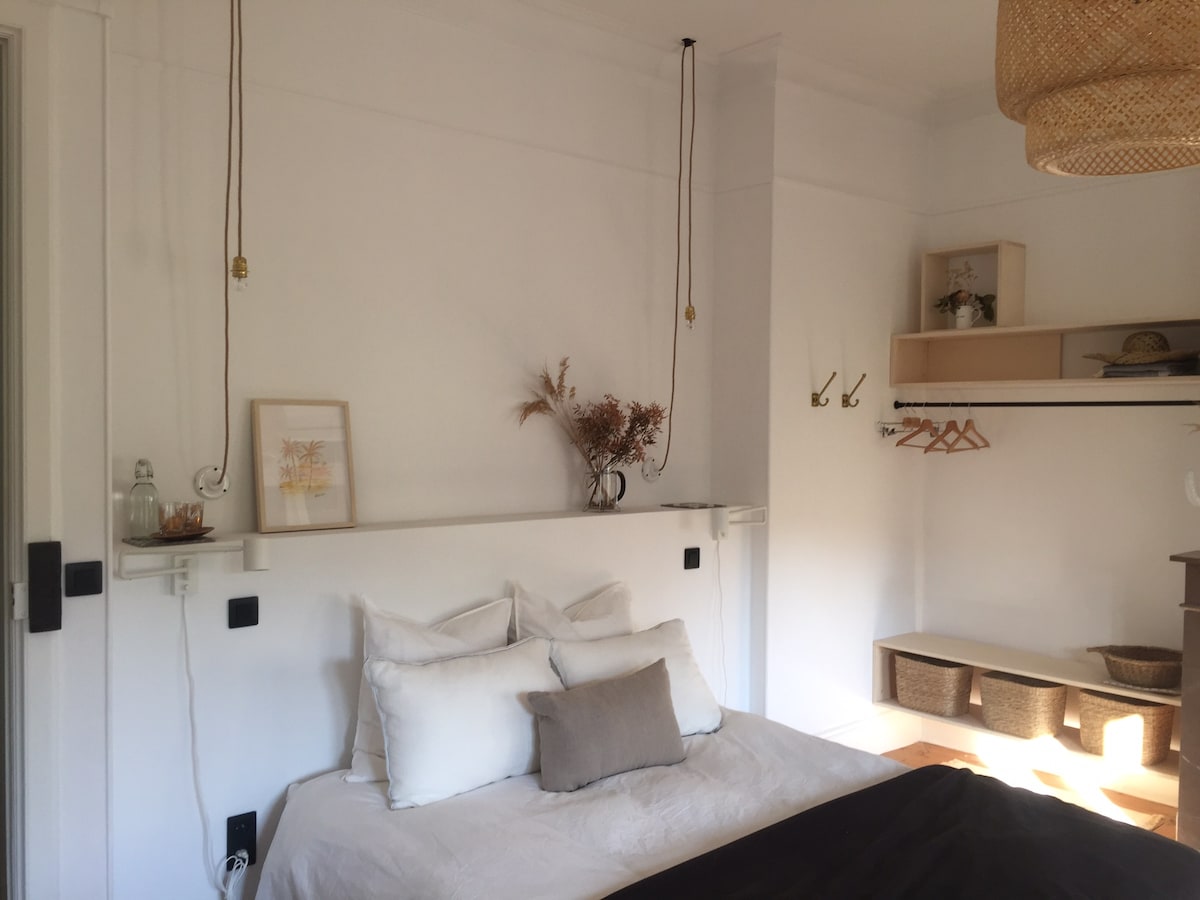 A spacious bedroom is presented with neutral white walls and soft natural light. A comfortable bed is adorned with a combination of white and taupe bedding. Shelving units showcase decorative items, and wall hooks are equipped for clothing storage. A woven pendant light adds texture.