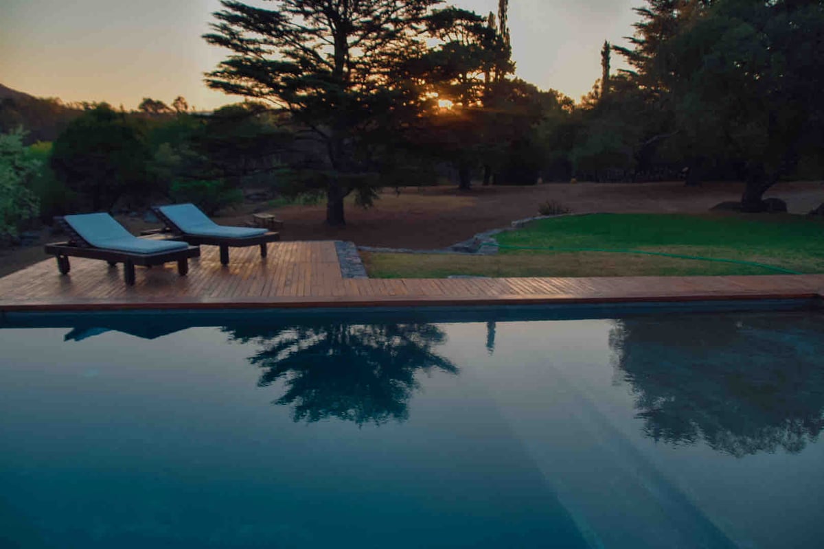 A serene outdoor area features a tranquil pool reflecting the surrounding trees and sunset. Two lounge chairs positioned on a wooden deck invite relaxation, while the lush green lawn complements the natural setting.