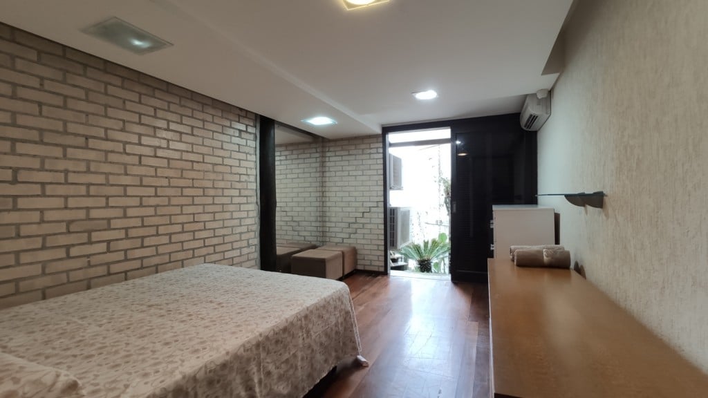 A spacious bedroom features a large bed covered with a patterned blanket, complemented by a wooden table and a mirror. Natural light enters through a glass door leading to a small outdoor area with plants, enhancing the overall brightness of the room.