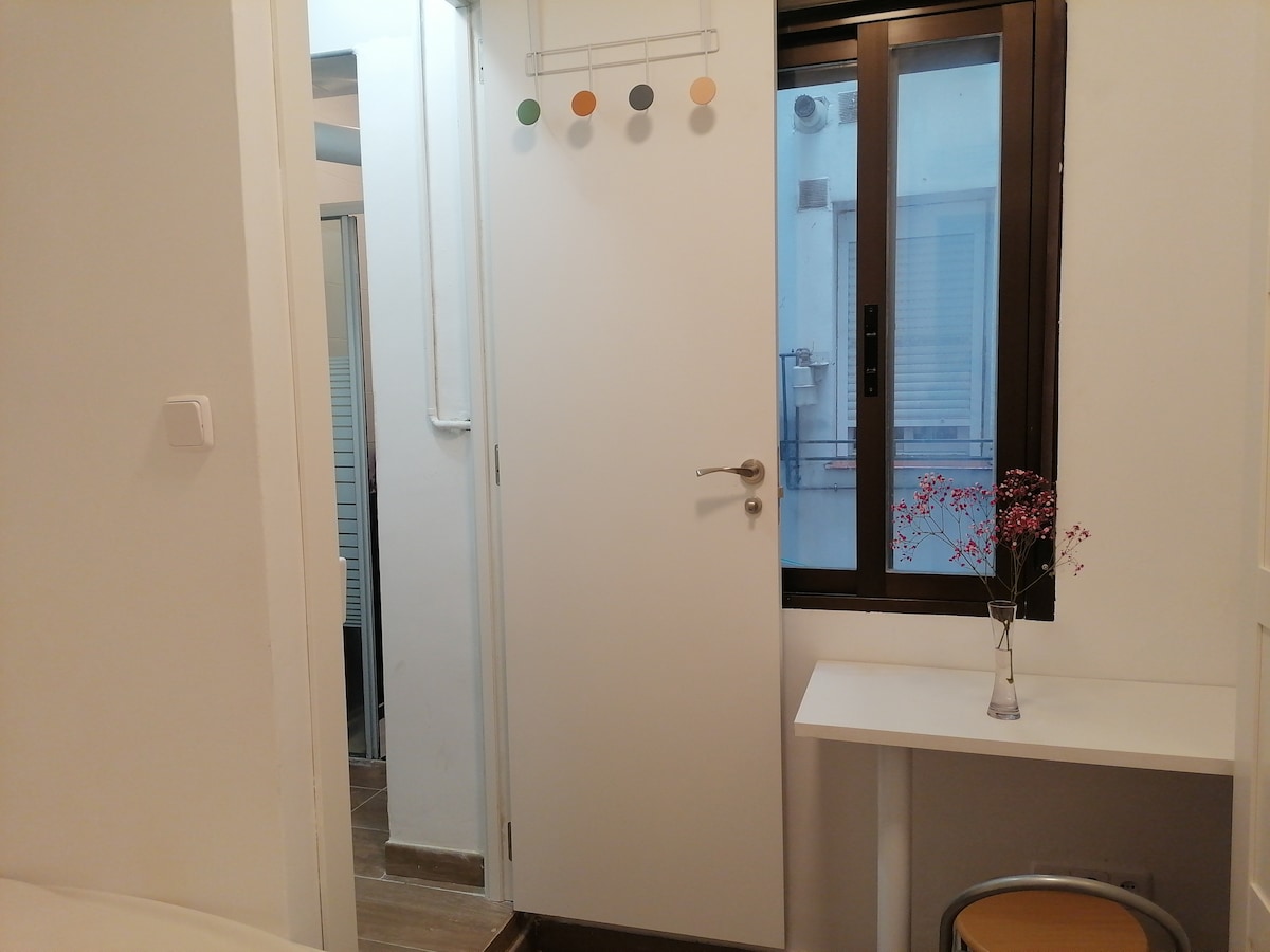 A well-lit room is showcased with a simple white door leading to another space. A small window allows natural light to enter, featuring a view of an exterior wall. A minimalist table sits beside a vase with dried flowers, adding a subtle accent.