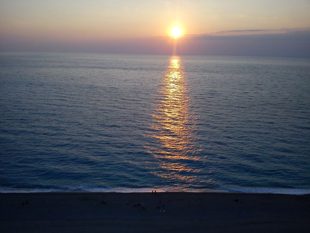 A sunset over a calm sea is captured, with golden reflections shimmering across the water. The horizon blends soft hues of orange and purple, while a shoreline is visible with a few figures along the beach, enjoying the peaceful scenery.