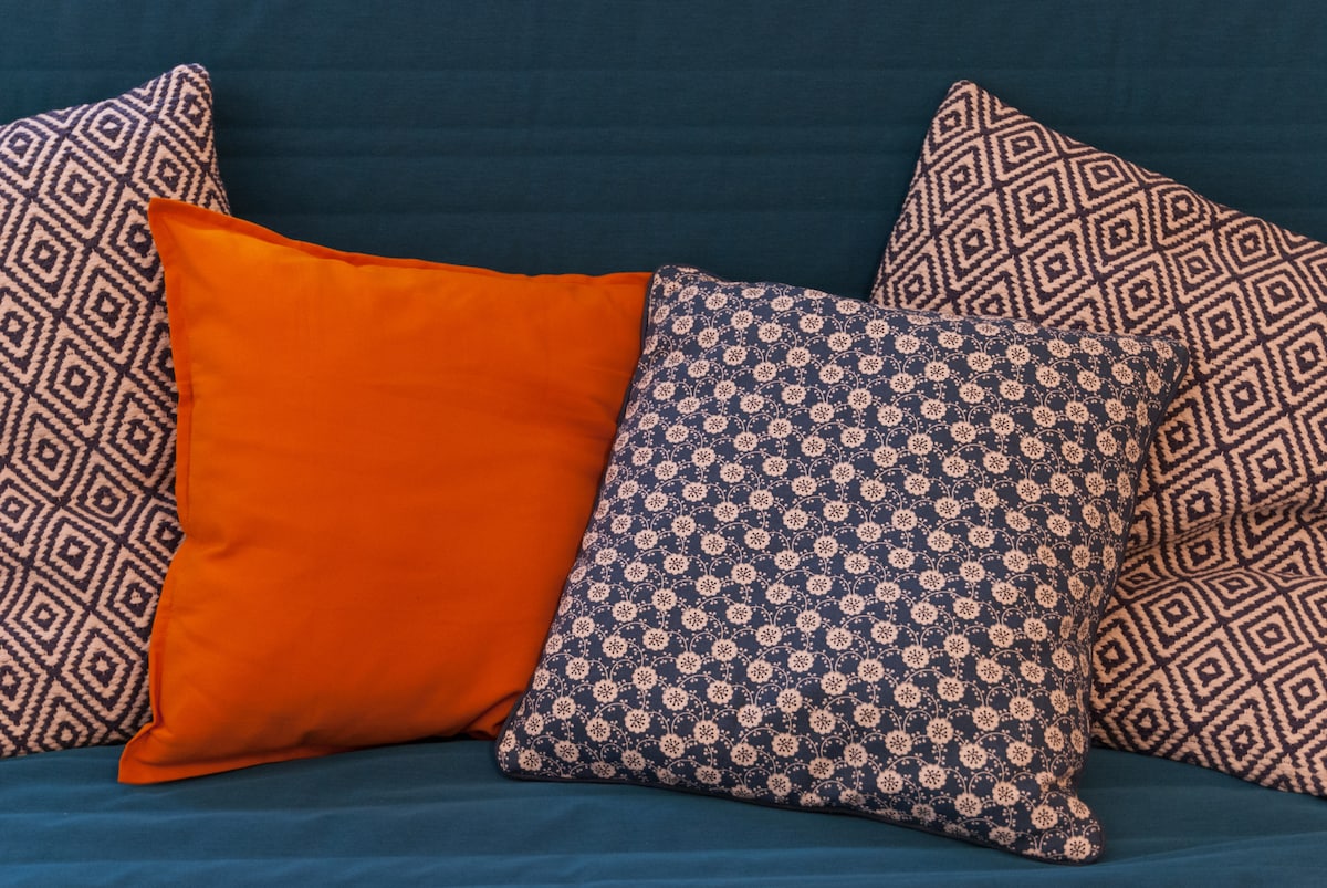 A cluster of decorative pillows is arranged on a sofa, featuring an orange pillow and two patterned pillows in navy blue and white. The background is a solid blue fabric, creating a cohesive and comfortable seating area.