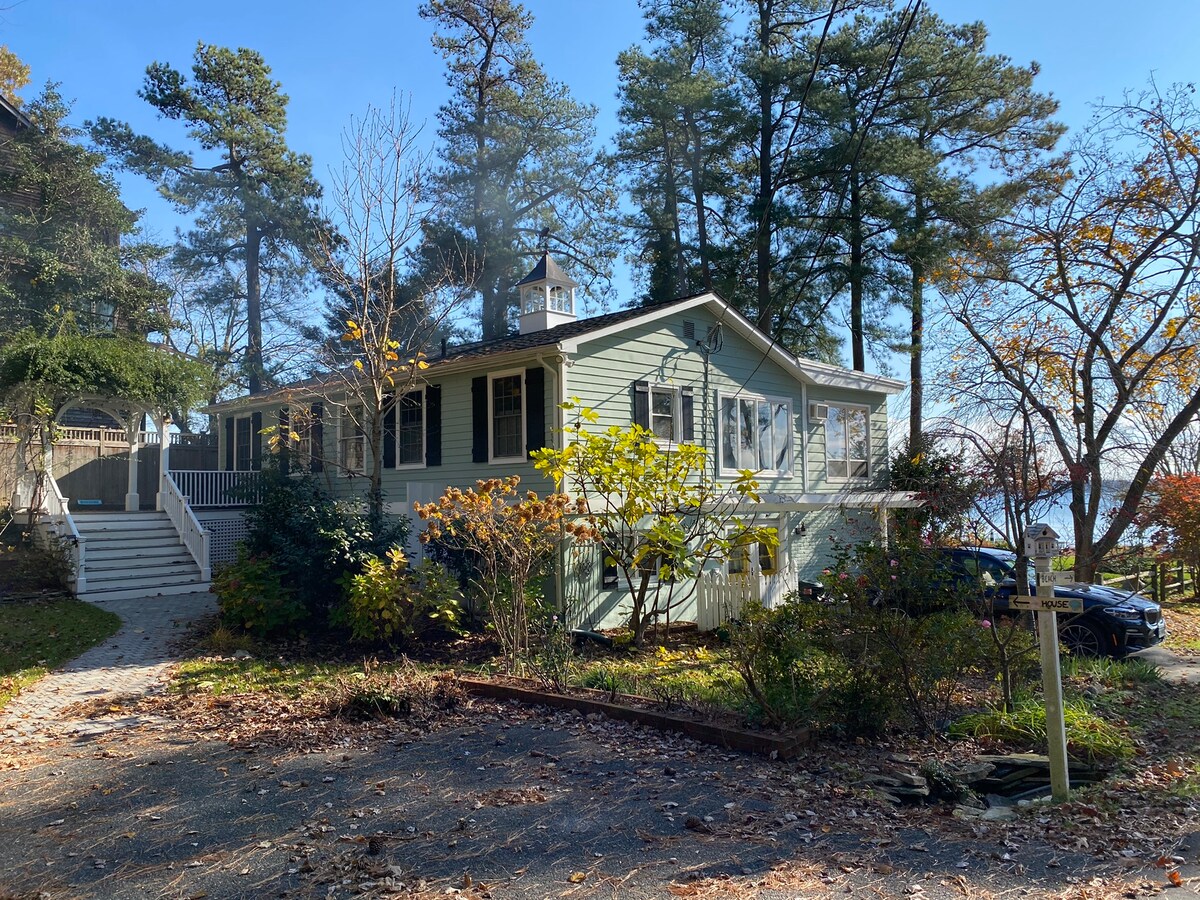 A charming bay house is set among mature trees, showcasing a light green exterior. Steps lead up to the welcoming entrance, while vibrant foliage frames the home's sides. A partial view of the bay is visible in the background, hinting at the serene surroundings.