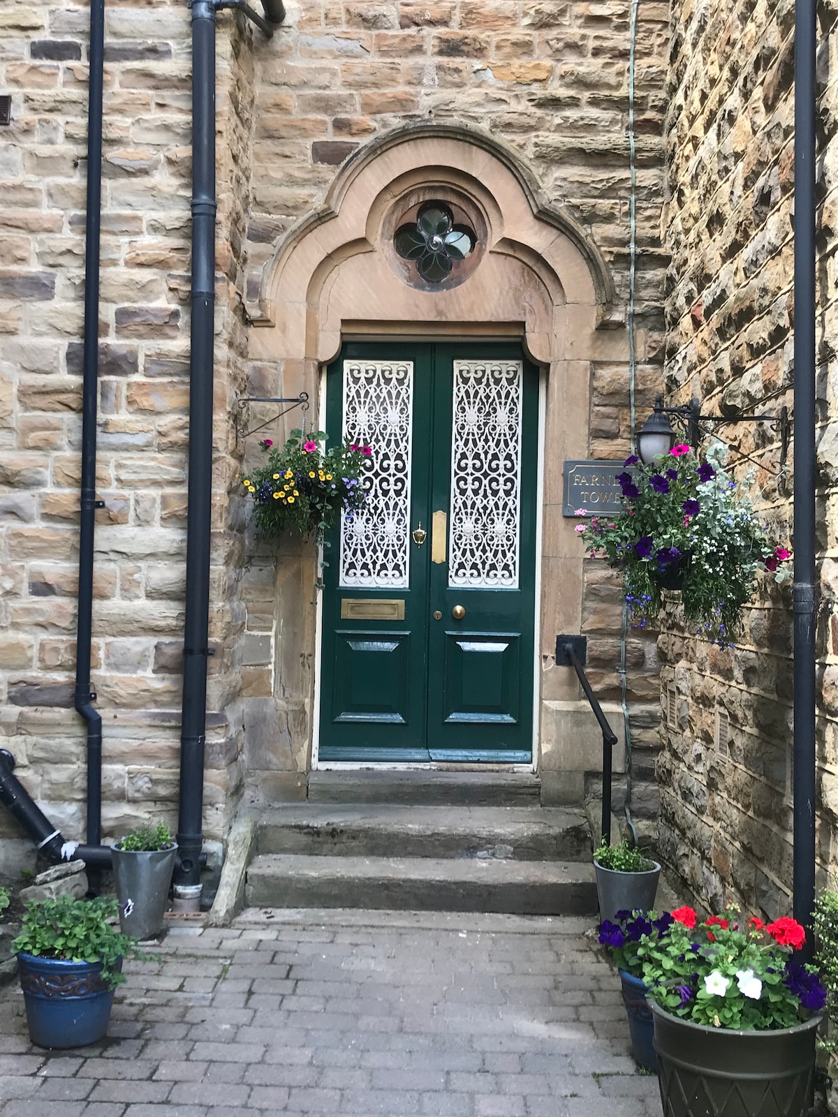 The entrance features a double green door with intricate lace detailing, set within a stone facade. Flower pots with colorful blooms are arranged along the steps, adding vibrancy to the entrance. A small stone pathway leads up to the door, creating a welcoming access.