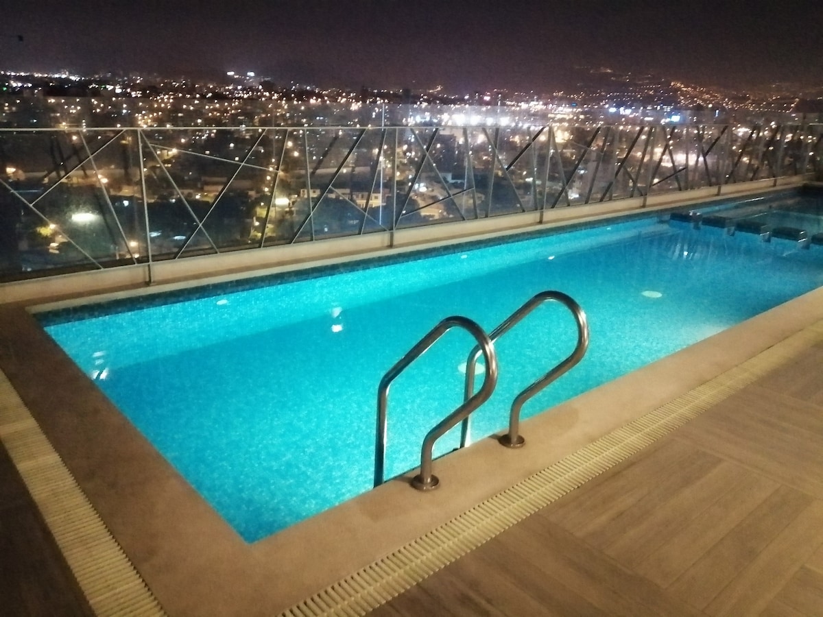 A rooftop pool glimmers under the night sky, surrounded by a glass railing that offers views of the city skyline. The pool features a set of steps for easy access, and the tranquil water surface reflects the nearby lights.