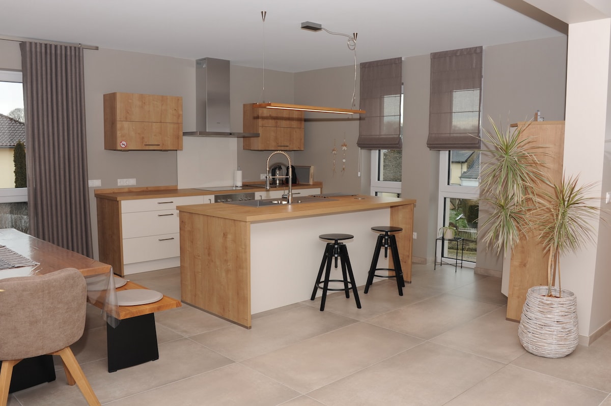 A modern open kitchen features wooden cabinetry and sleek appliances. A central island provides ample workspace, complemented by bar stools. Natural light enters through large windows, while minimalist decor, including potted plants, enhances the inviting atmosphere. A dining area with a wood table is visible.
