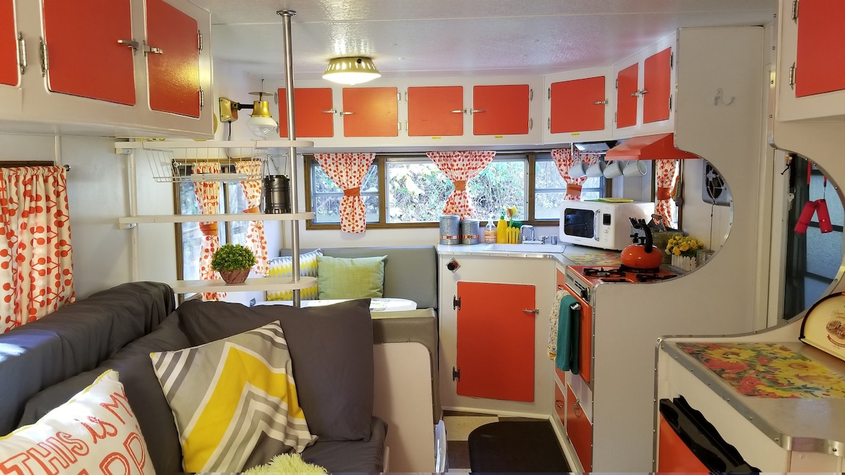 The interior of the vintage camper features a living area with a cozy sofa adorned with patterned cushions. Bright orange cabinetry complements the white walls, while colorful curtains frame the windows. A small kitchen area includes essential appliances, with a dining nook visible in the background.