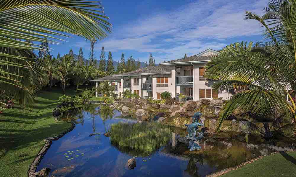 A tranquil resort building is surrounded by lush greenery and palm trees, situated near a serene pond that reflects the structure. The landscape features various rocks and a decorative statue, enhancing the natural beauty of the environment.