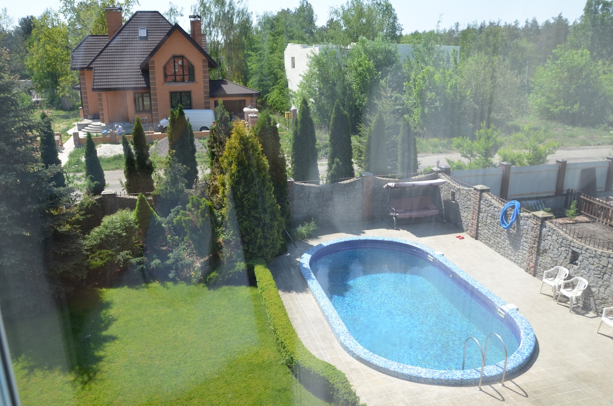 A view from above captures a rectangular pool framed by manicured greenery and patio space. Surrounding trees provide privacy. The crystal-clear water reflects sunlight, while the pool area features several white lounge chairs arranged nearby.