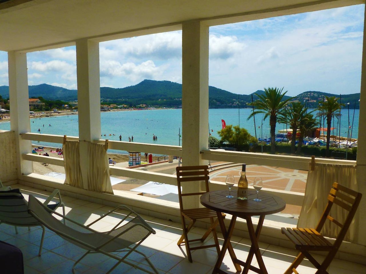 A spacious terrace offers a view of the beach and harbor, framed by palm trees and distant hills. Two lounge chairs and a small table set on the terrace invite relaxation. Natural light fills the area through wide openings, connecting the indoors with the outdoor scenery.