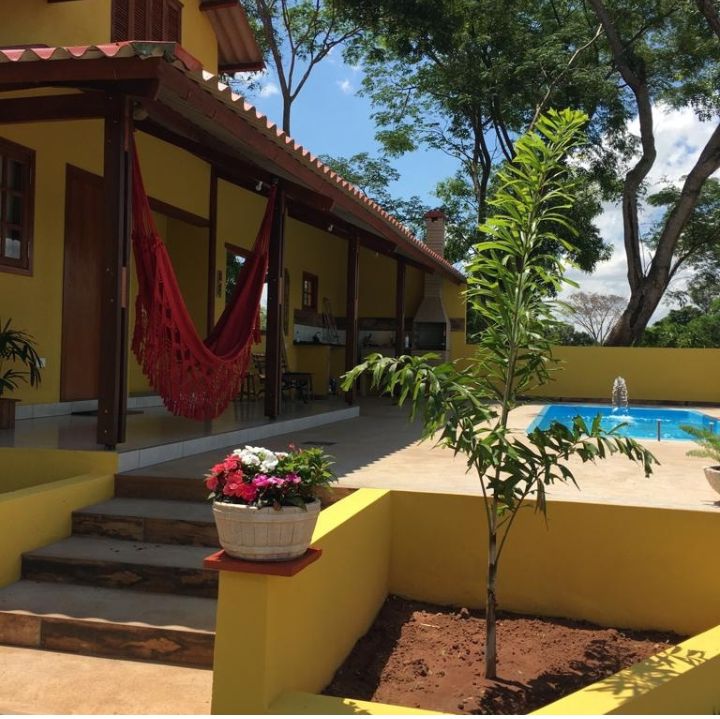 A vibrant yellow house features a spacious porch with a red hammock. Lush greenery surrounds the area, complementing a small tree and flower arrangement. A sparkling blue pool can be seen in the background, reflecting the sunlight and inviting relaxation.