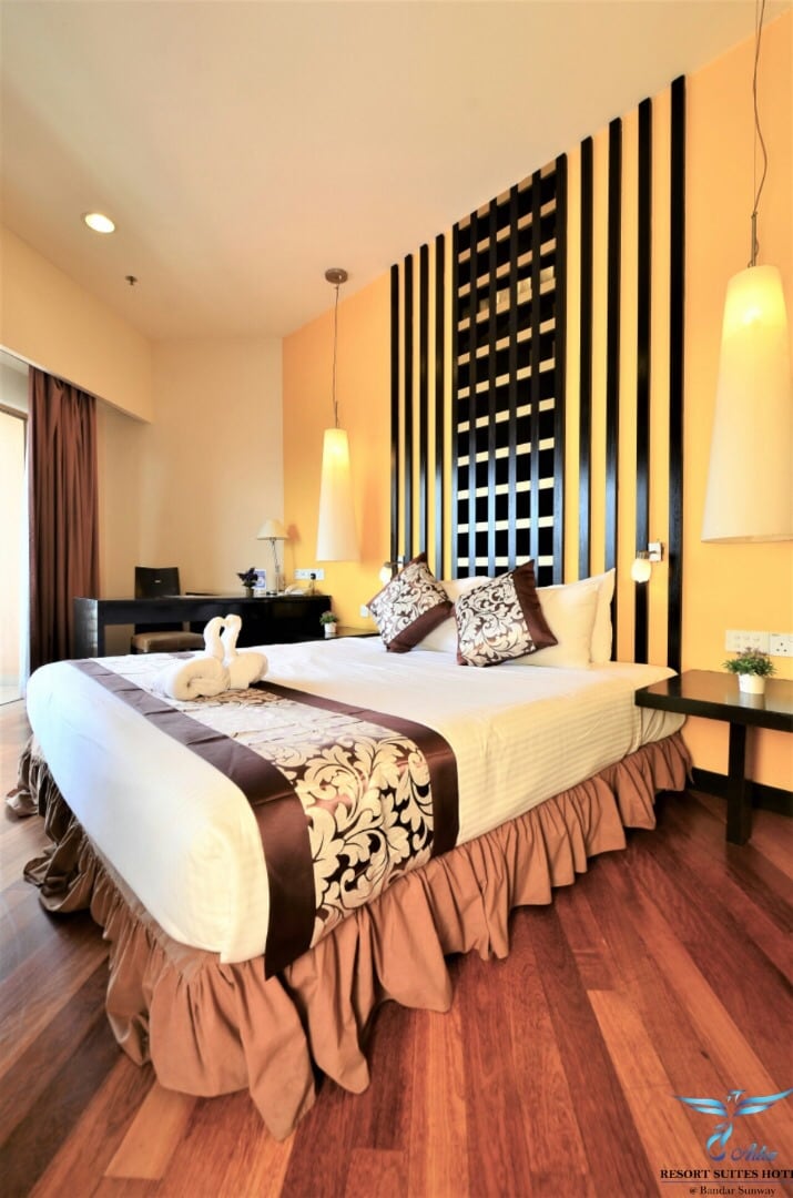 The room features a spacious bed adorned with decorative pillows and a stylish bed skirt. A work desk is positioned nearby, illuminated by pendant lights. Natural light enters through a glass door leading to a balcony, enhancing the warm wooden flooring.