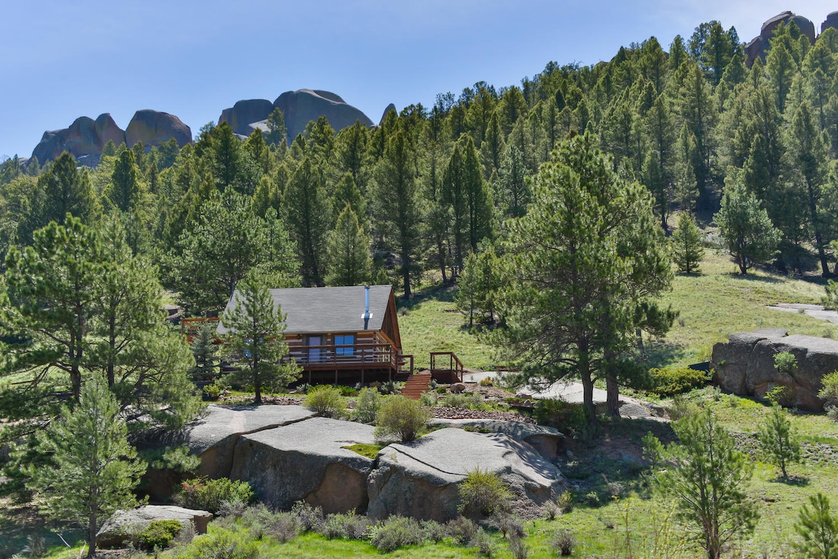 The rustic cabin is nestled among towering pine trees, surrounded by sprawling natural landscapes. Rocky outcrops and vibrant greenery complement the serene setting, while the cabin's wooden exterior harmonizes with the environment.