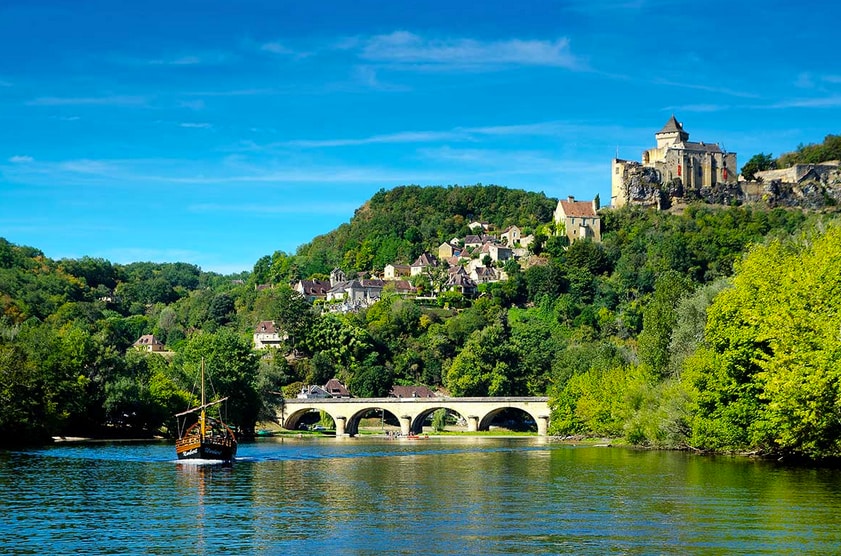 A serene landscape features a river flowing gently in the foreground, with a historical castle perched on a hillside. The surrounding area is dotted with quaint houses and lush greenery, while a wooden boat navigates the water, adding a sense of tranquility to the scene.