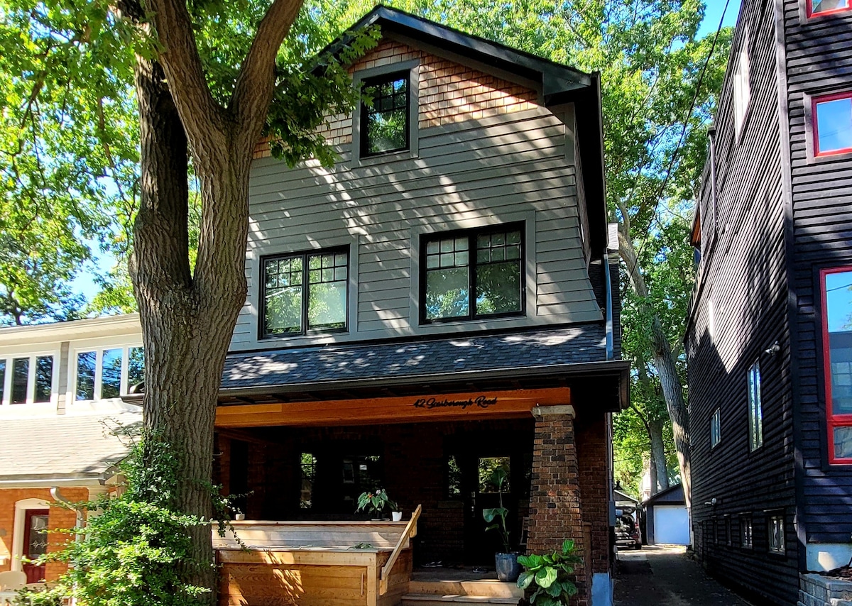 The exterior of a multi-story home is visible, featuring wood and brick construction with large, square windows. A wooden porch extends across the front, adorned with potted plants. Surrounding trees provide shade, and neighboring structures can be seen on either side.