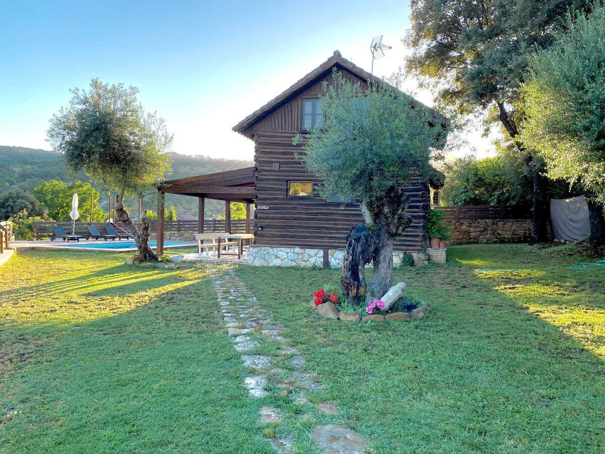 A charming wooden house is set in a lush green landscape, surrounded by trees and flowering plants. A stone pathway leads to the front of the house, with a pool visible in the background. The structure features a rustic design and is bathed in warm sunlight.