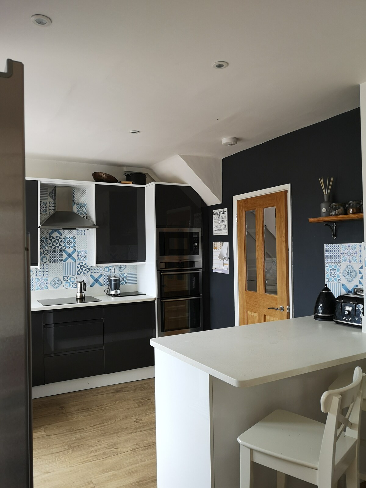 The kitchen features modern cabinetry in contrasting dark and light tones, complemented by patterned backsplash tiles. A breakfast bar with white stools is present, and stainless steel appliances are neatly arranged, creating an organized and functional cooking space.