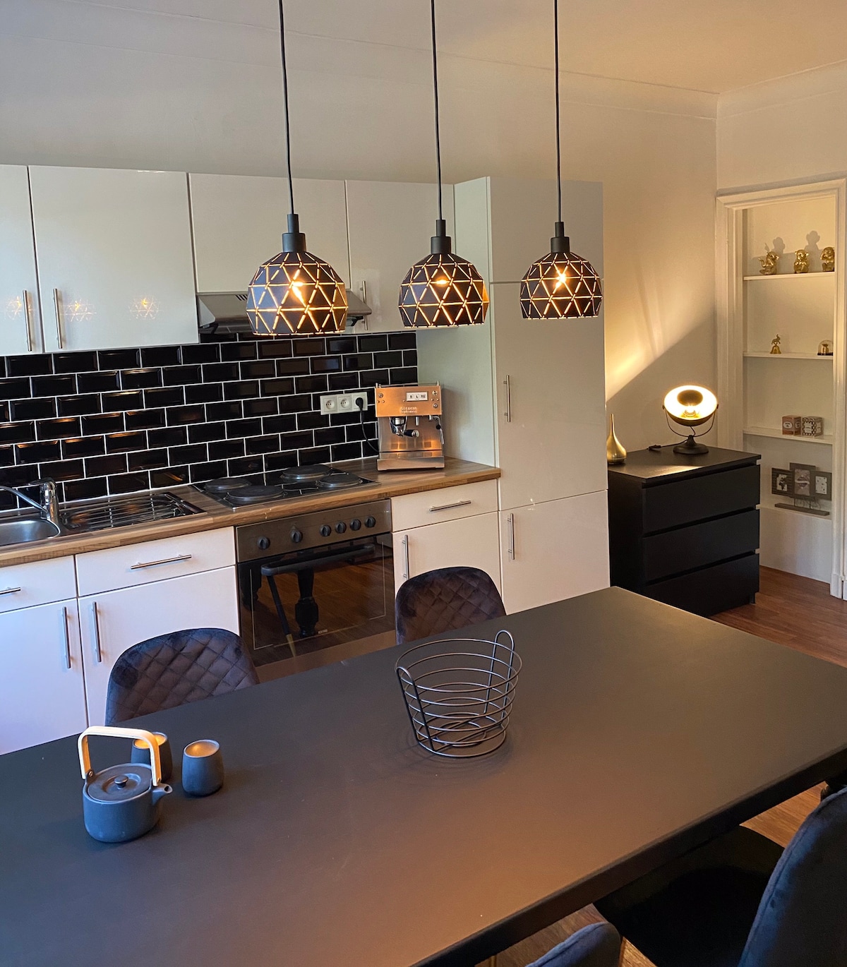 A modern kitchen space is presented, featuring sleek white cabinets and a black tile backsplash. Three pendant lights hang above a dark dining table, complemented by stylish black chairs. A coffee machine and various kitchen appliances are neatly arranged on the countertop.