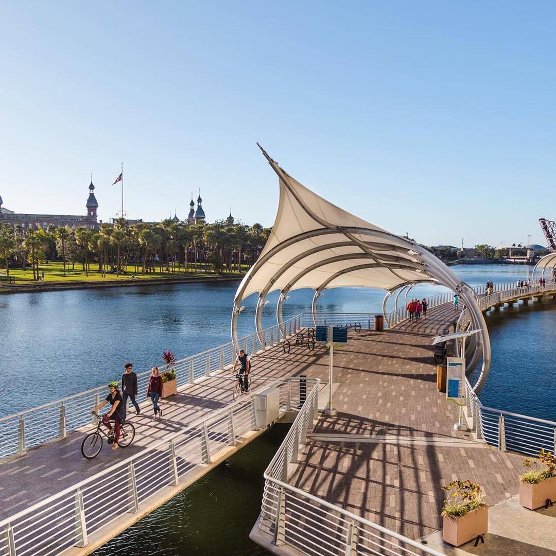 A scenic waterfront walkway extends over calm waters, featuring a distinctive white canopy structure. People can be seen walking and biking along the path. Lush palm trees line the shore, with historic buildings visible in the background against a clear blue sky.