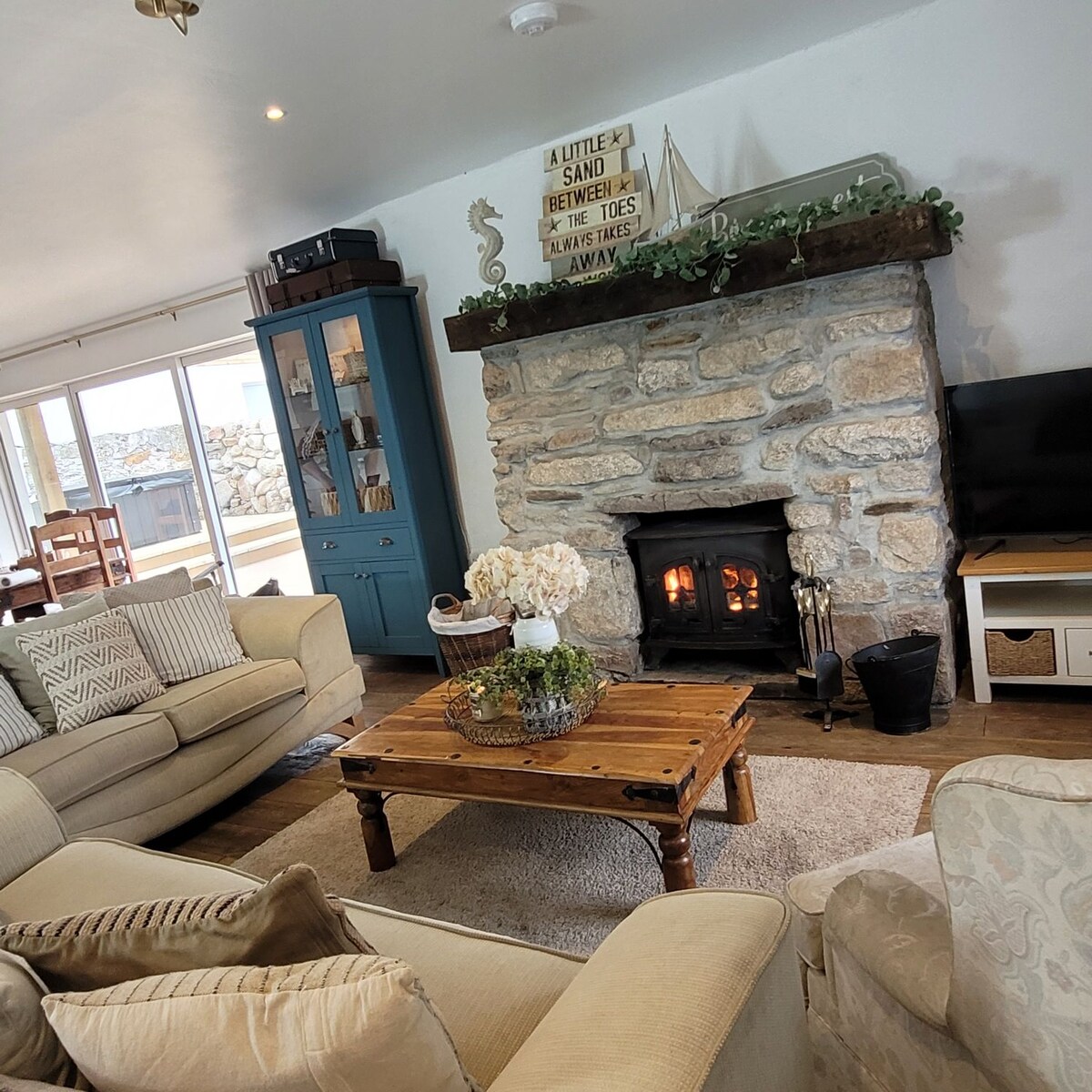 A cozy living area features soft furniture arranged around a rustic wooden coffee table. A stone fireplace provides warmth, with a flickering flame visible inside. Light-colored walls and a blue cabinet enhance the inviting ambiance, while natural light streams through large windows.