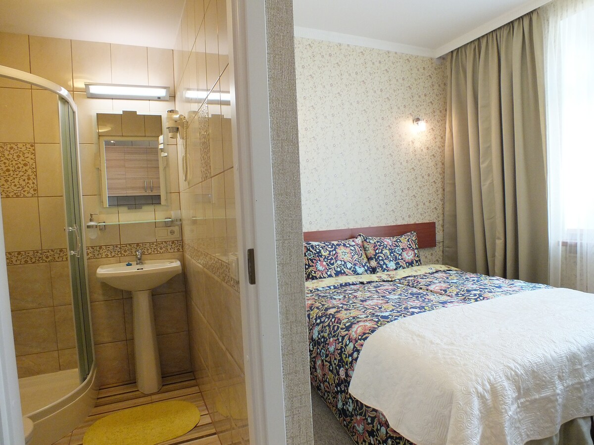 A small studio room features a spacious bed adorned with floral bedding. A separate en-suite bathroom is visible, equipped with a shower, sink, and a well-lit mirror. The room is enhanced by natural light streaming through a nearby window, complemented by soft curtains.