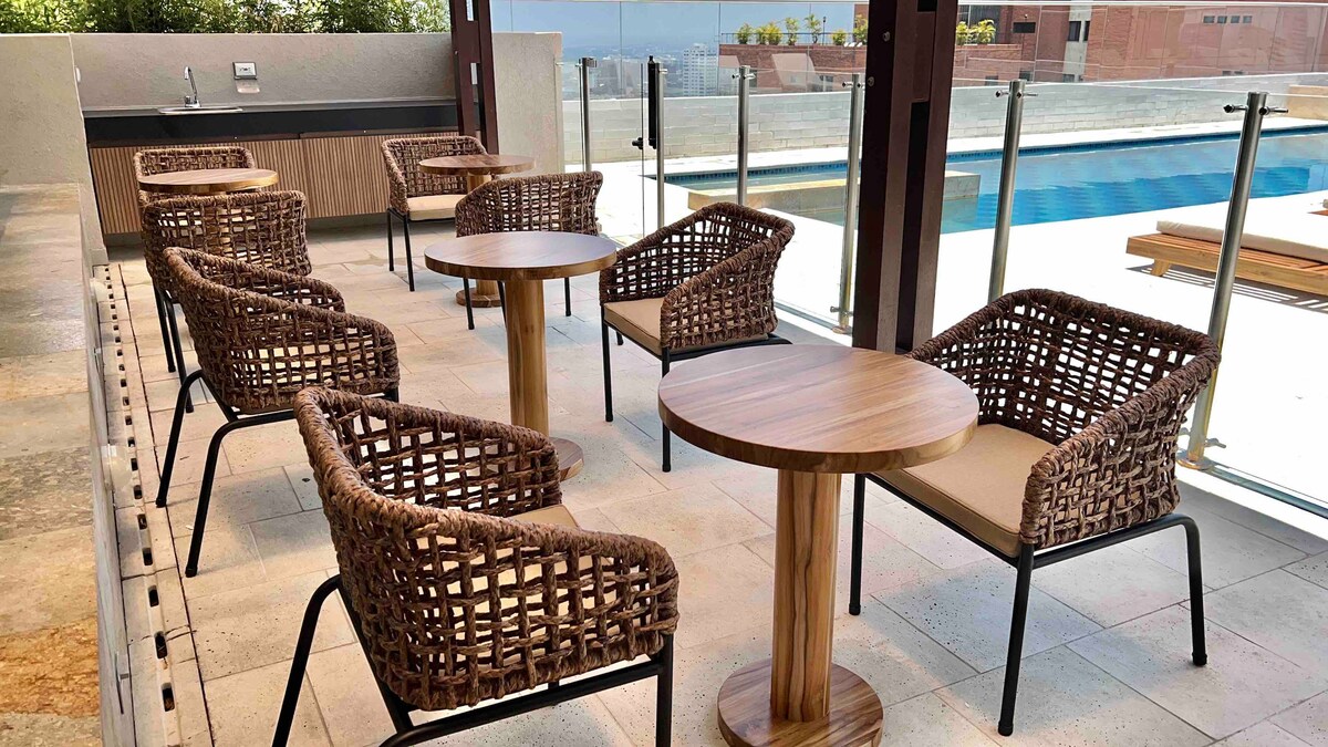 An outdoor lounge area features six woven chairs paired with two circular wooden tables. The space is set against a backdrop of a swimming pool and has glass railings providing an open view, complemented by natural elements and soft lighting.