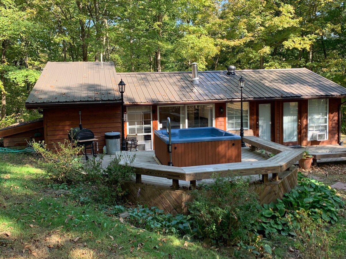 The cabin's exterior showcases a wooden facade surrounded by trees, with a deck leading to a hot tub. Large windows provide views of the forest, while a charcoal grill and outdoor seating are positioned nearby. A gentle slope enhances the secluded ambiance.