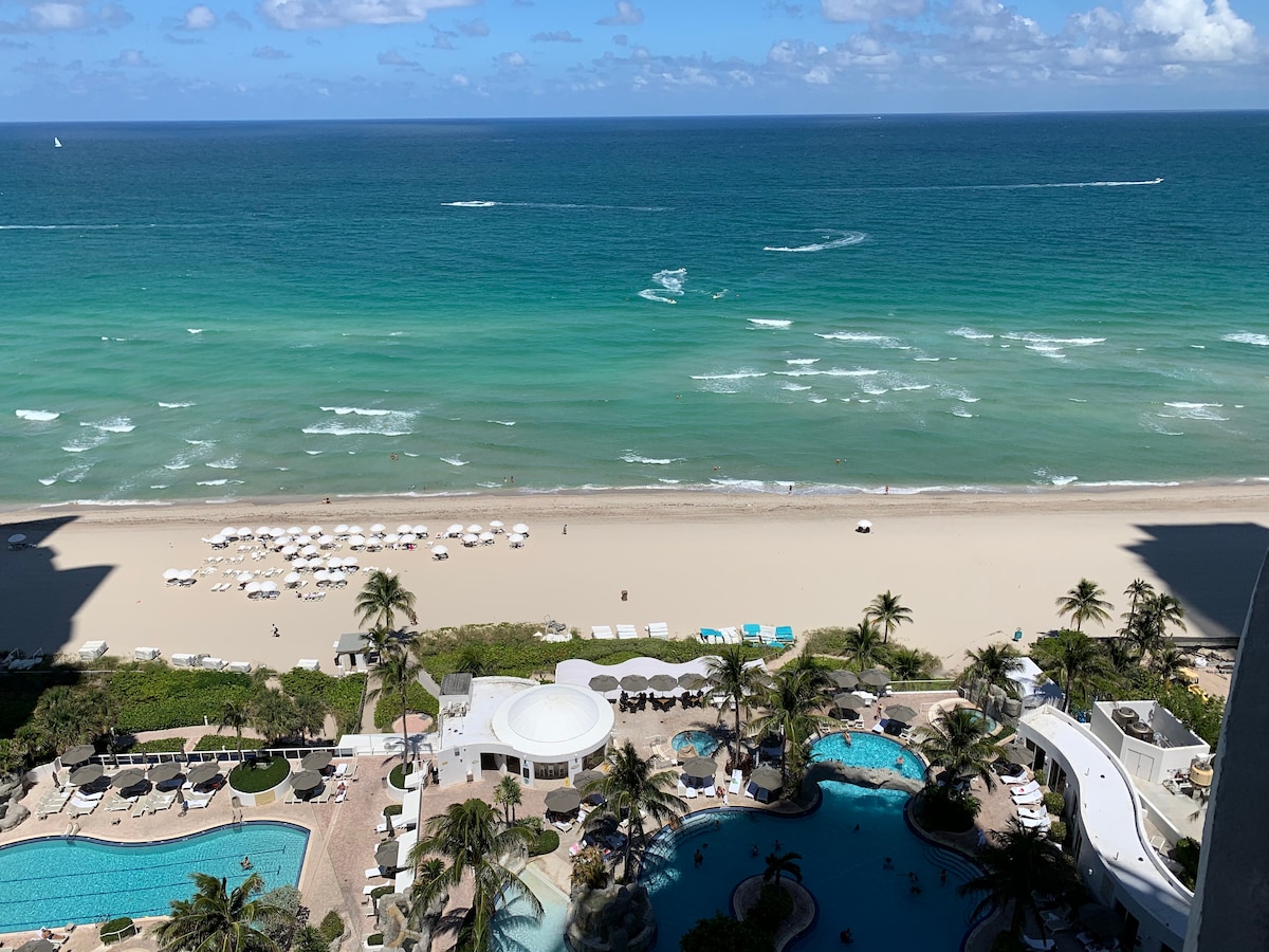 A direct ocean view captures turquoise waters and a sandy beach lined with white umbrellas. Below, the resort's pools, surrounded by palm trees, create an inviting setting. Multiple lounge chairs and cabanas are visible, enhancing the tranquil atmosphere of this beachfront location.