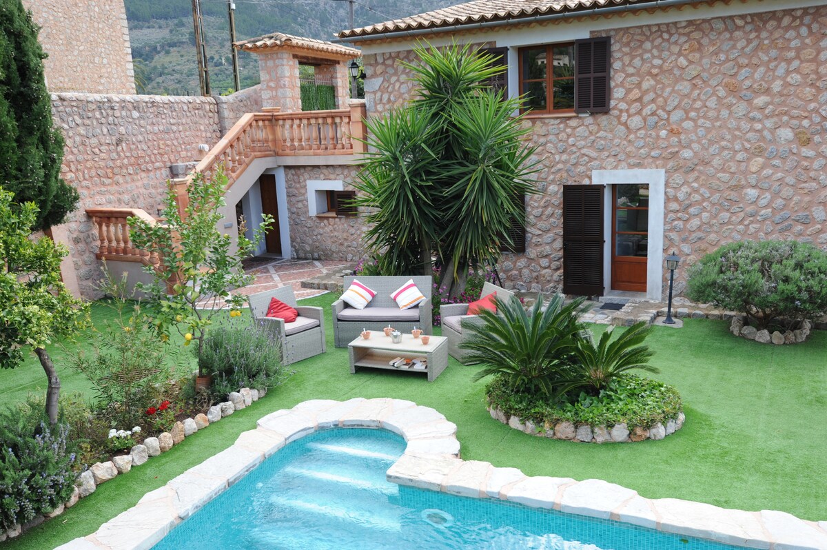 A stone house features a lush garden with various plants and a sparkling pool. Comfortable seating is arranged around a low table. A staircase leads to the upper level, while a doorway opens into the interior of the home.