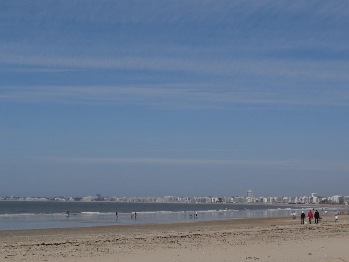 A sandy beach stretches along the coastline, with gentle waves lapping at the shore. Visitors are seen strolling along the water's edge, while the skyline of a coastal town is visible in the background under a clear blue sky.