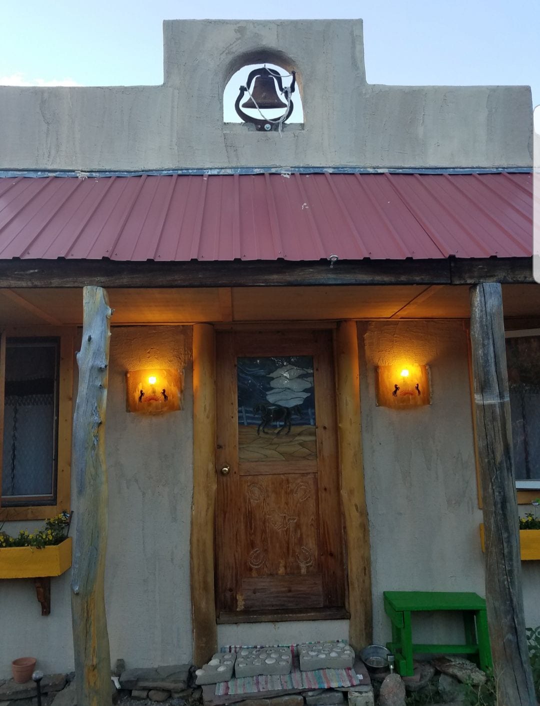 The rustic cabin entrance features a wooden door adorned with a colorful stained glass design. Two warm lights cast a soft glow on either side. A decorative bell hangs above the door, and the entrance is framed by natural wooden beams and a corrugated metal roof.