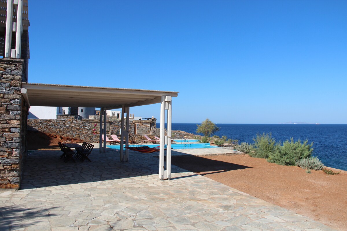 A shaded patio area is shown with wooden lounge chairs and hammocks positioned to overlook the sea. The stone path leads to a swimming pool bordered by greenery, while a clear blue sky is visible above, enhancing the sense of outdoor relaxation.