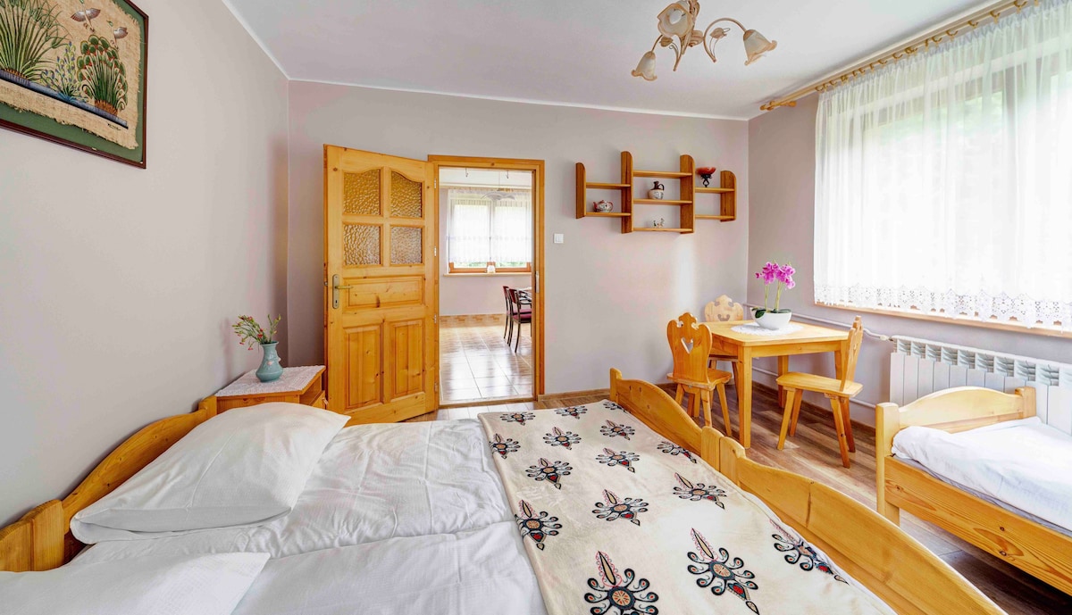 A cozy bedroom features a wooden double bed adorned with a decorative blanket. A small dining area is visible, equipped with a table and two chairs, accompanied by a single bed. Natural light filters through the large window, enhancing the space's warmth.