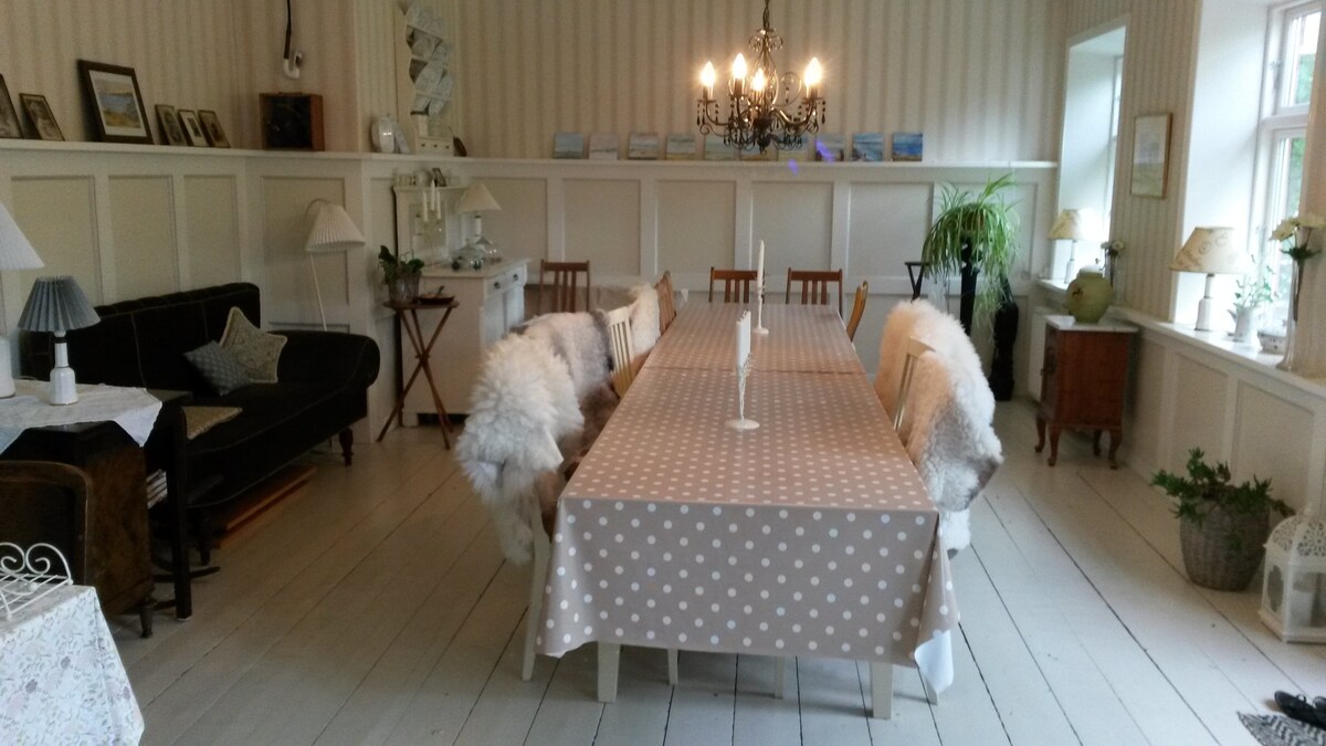 A spacious dining area features a long table covered with a polka-dot tablecloth, surrounded by several wooden chairs, some draped with cozy throws. A chandelier hangs above, illuminating the space, which is adorned with plants and wall decor, creating a welcoming atmosphere.