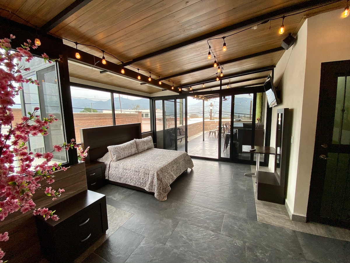 A bright sleeping area features a spacious bed with a floral patterned blanket. Glass walls provide views of the outdoor terrace, while warm string lights illuminate the ceiling. A dark wooden nightstand complements the aesthetic, and a television is mounted on the wall.