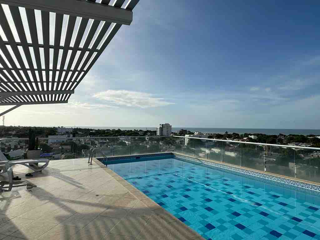 The rooftop pool features a clean, modern design with a glass barrier providing unobstructed views of the ocean and surrounding city. Sun loungers are placed nearby, and the sun casts gentle shadows across the tiled deck under a bright blue sky.