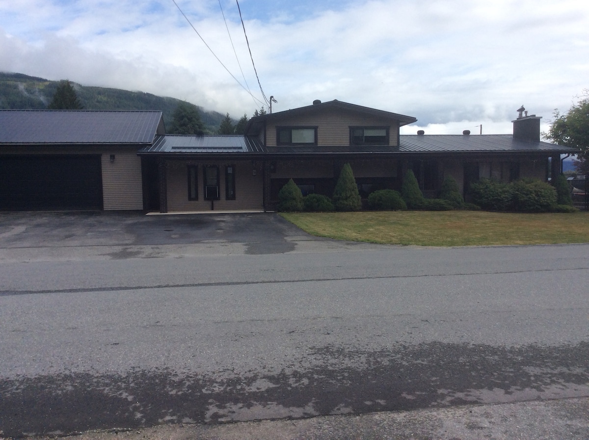 A spacious two-story building features a mix of stone and wood siding under a metal roof. The front entrance is accessible, flanked by manicured bushes and a large lawn. Power lines run overhead, and a gravel driveway is visible, leading to a garage on the left side.